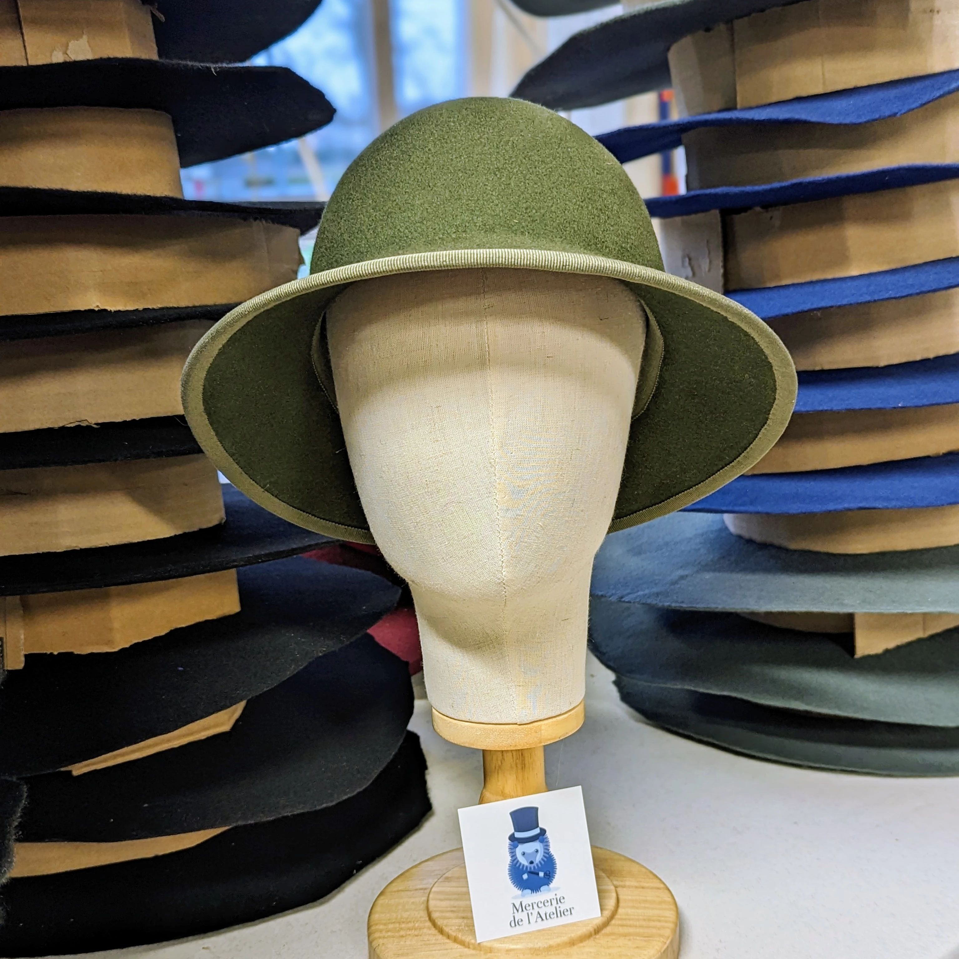 Chapeau en feutre vert olive sur une forme de t&ecirc;te, entour&eacute; de formes &agrave; chapeau en bois dans l'atelier